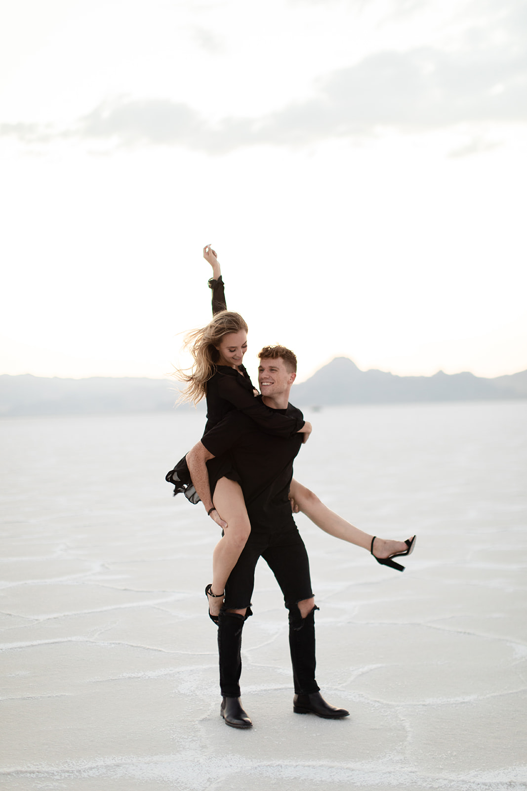 Utah engagement session at the salt flats with a cute playful bride-to-be piggybacking on her fiance dress in a black flowy dress and tall black heels. Utah wedding photography utah engagement photography session salt flats engagement session engagement session outfit inspiration what to wear for an engagement session playful photography sessions in norrthern utah #utahbrideandgroom #destinationproposal #destinationengagement #destinationengagementphotography #engagementoutfit #engagementphotographytips #utahweddingphotography #dcweddingphotography #virginiaweddingphotography #virginiaengagementsession #engagementsessiontips