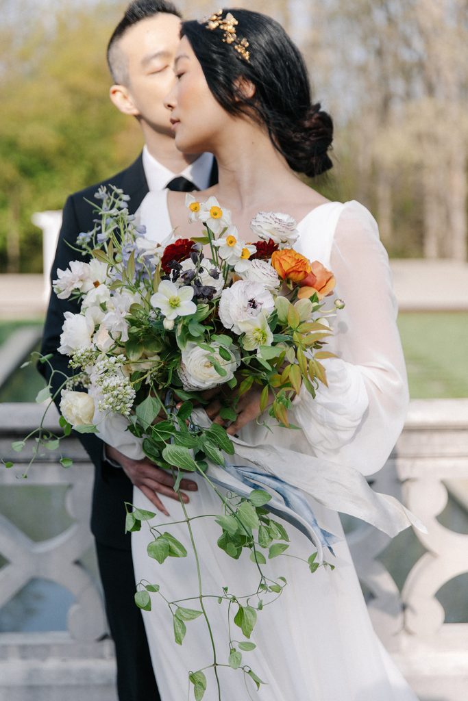 Planning a Villa Wedding: 9 Do's and Don'ts. Groom hugs bride from behind and kisses her cheek as she looks back at him while carrying her wedding bouquet