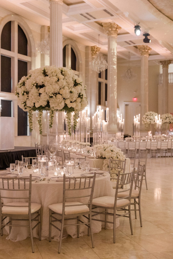 The wedding reception venue with tall windows, white walls and a huge white rose bouquet as the table centerpiece