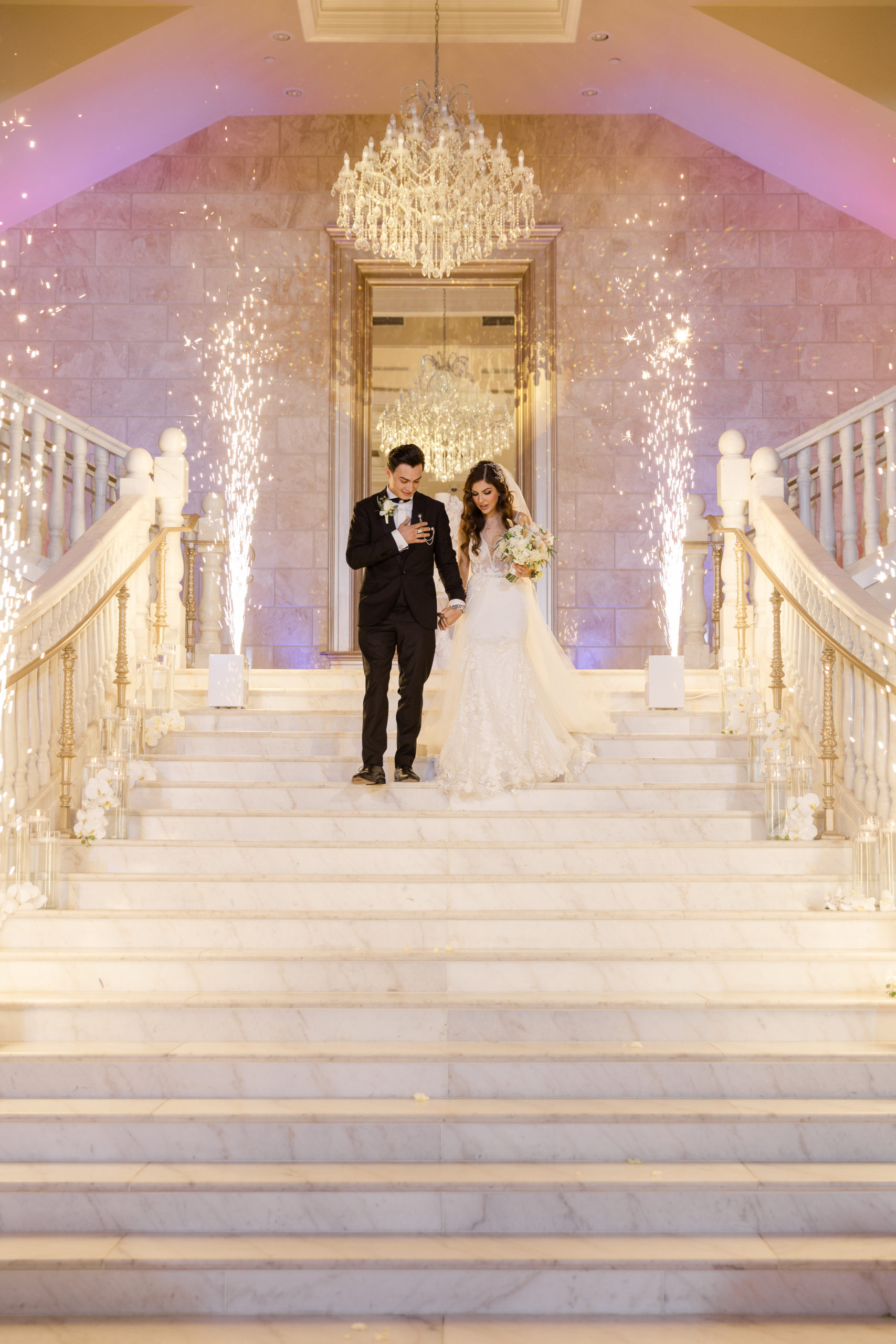 Bride and groom holding hands as they descend down the luxurious walkway to their ceremony location wth a chandelier above and fountain fireworks