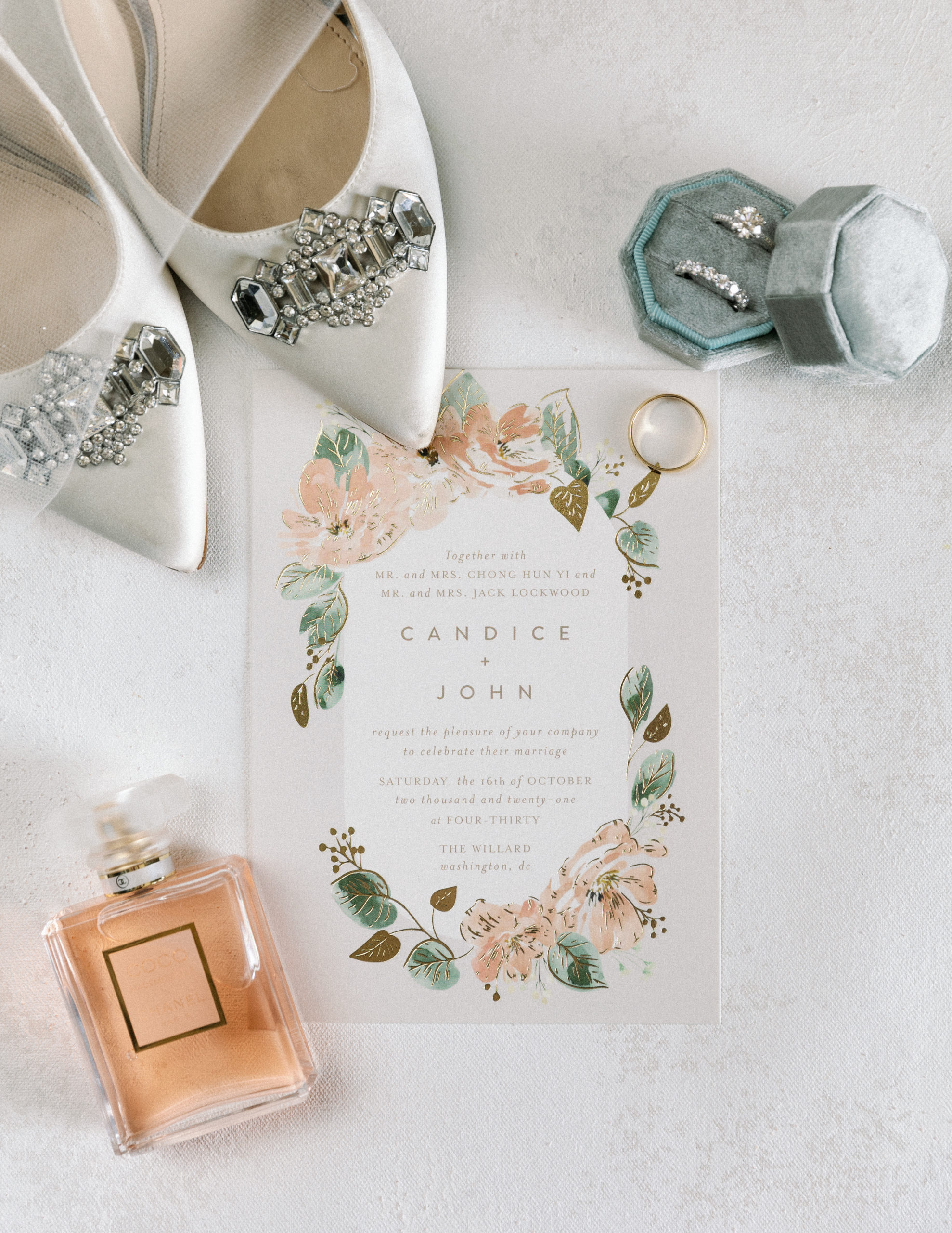 Flat-lay photo of the wedding invitation with artistic floral illustrations laid with a perfume bottle, wedding rings, and the bride's wedding shoes
