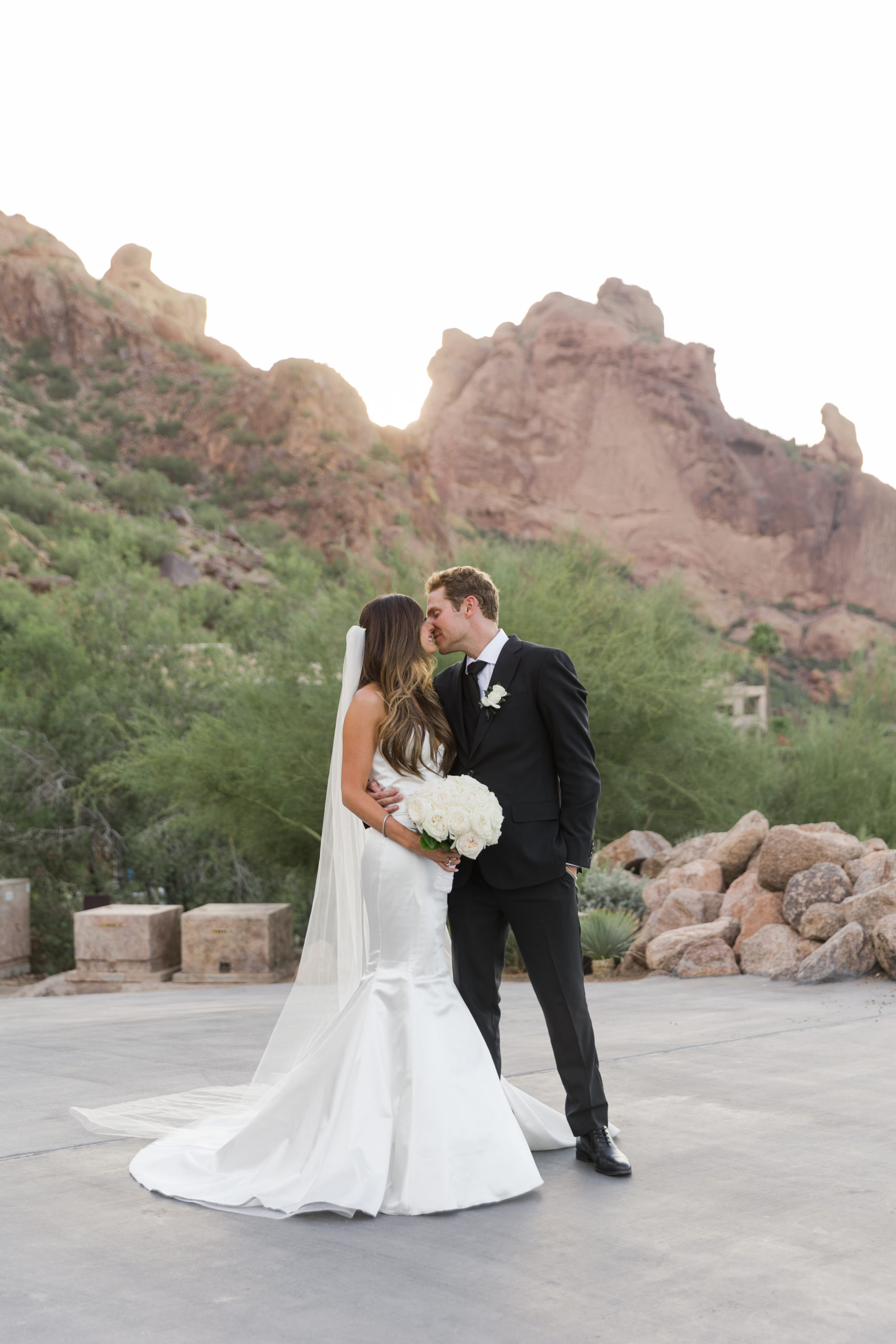 Groom leans in to kiss the bride as she smiles and holds on to her bridal bouquet during their elegant Camelback Mountain Wedding, captured by Kyla Jeanette