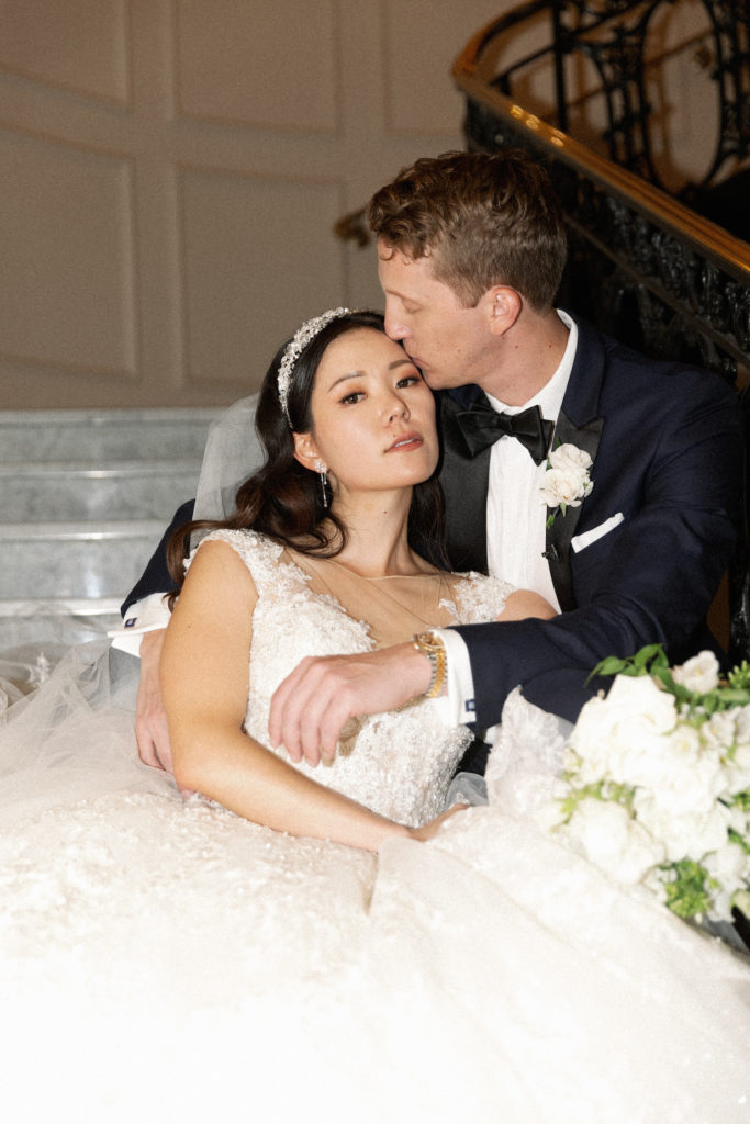 Groom plants a kiss on the bride's forehead while she looks enchantingly at the camera during their chic Willard Hotel wedding shoot with Kyla Jeanette Photography