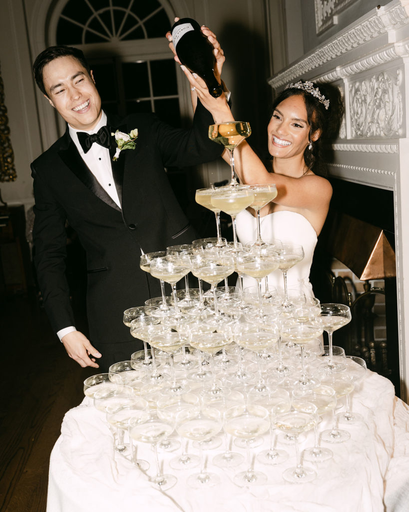 Bride and groom smile as they pour champagne over glass tower captured by elegant wedding photographer Kyla Jeanette